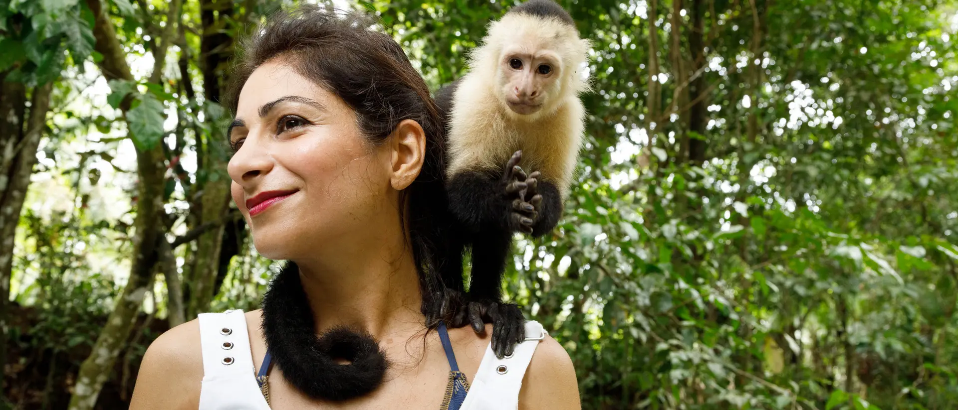 A woman smiles while a capuchin monkey perches on her shoulder in a lush green forest.