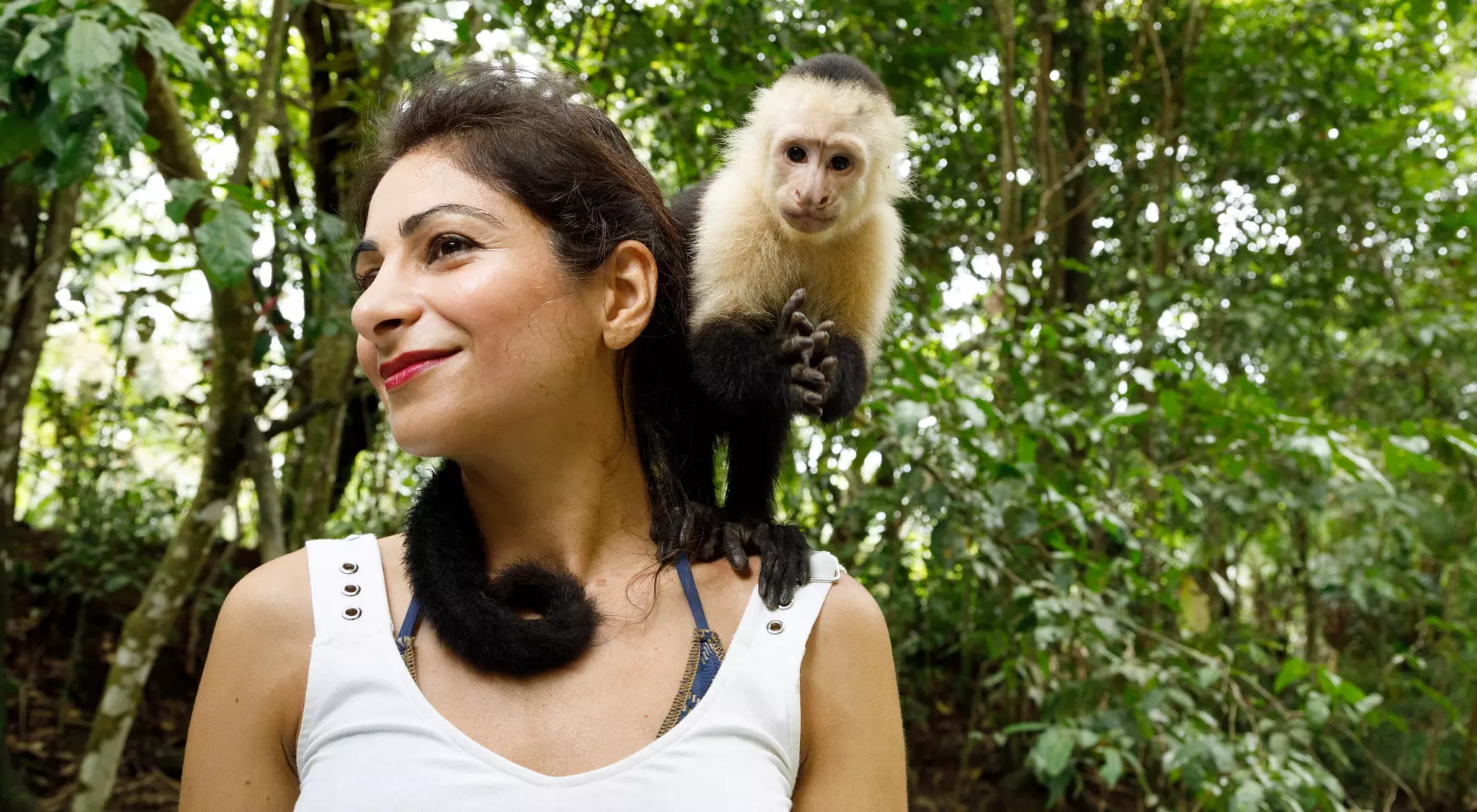 A woman smiles while a capuchin monkey perches on her shoulder in a lush green forest.