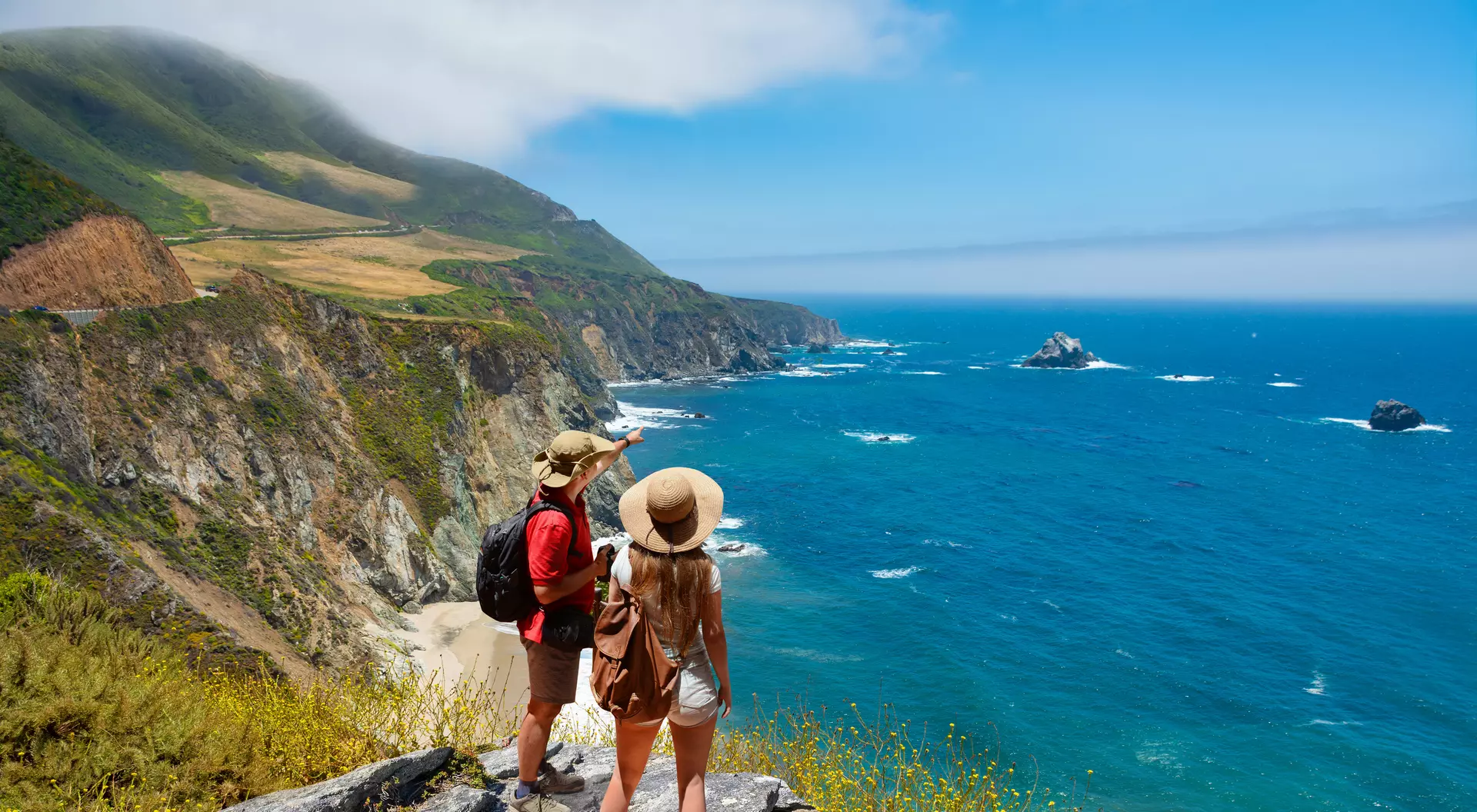 Two people stand on a rocky cliff, gazing at a scenic ocean view with cliffs and waves under a clear blue sky.