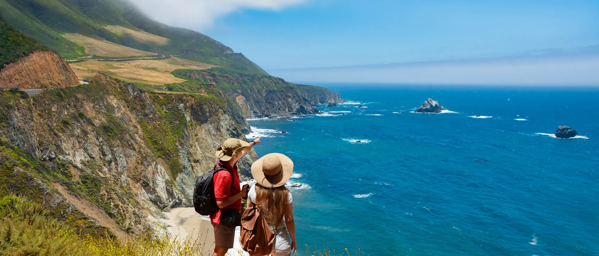 Two people stand on a rocky cliff, gazing at a scenic ocean view with cliffs and waves under a clear blue sky.