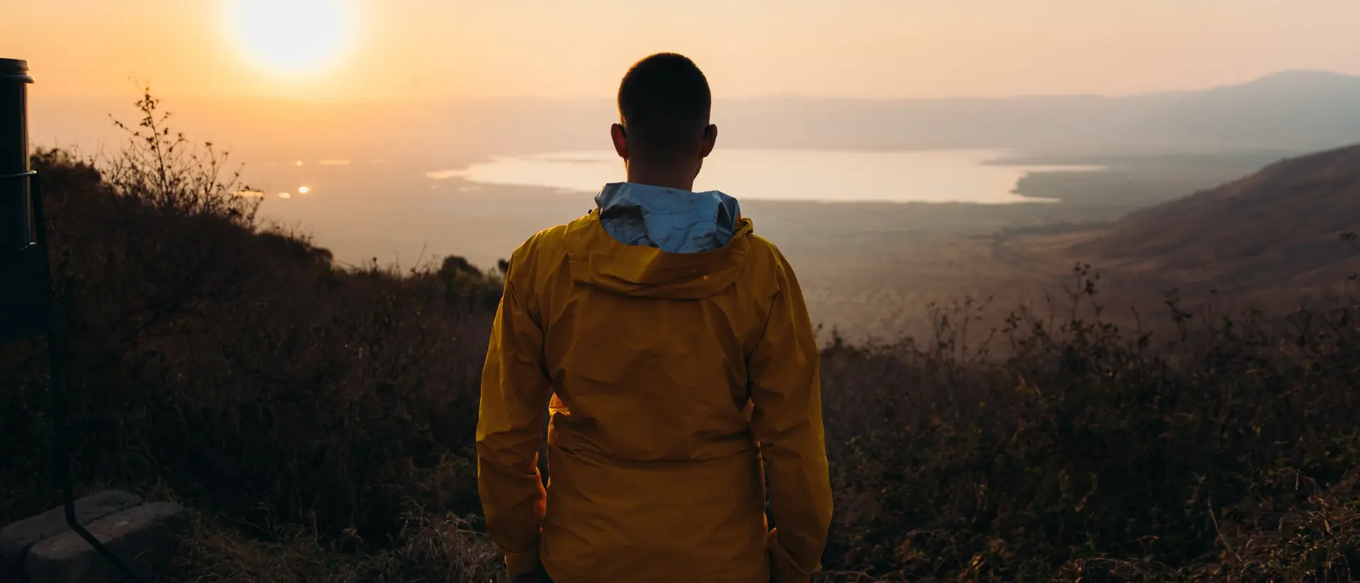 A person in a yellow jacket stands facing a sunset over a lake, surrounded by hills and vegetation.