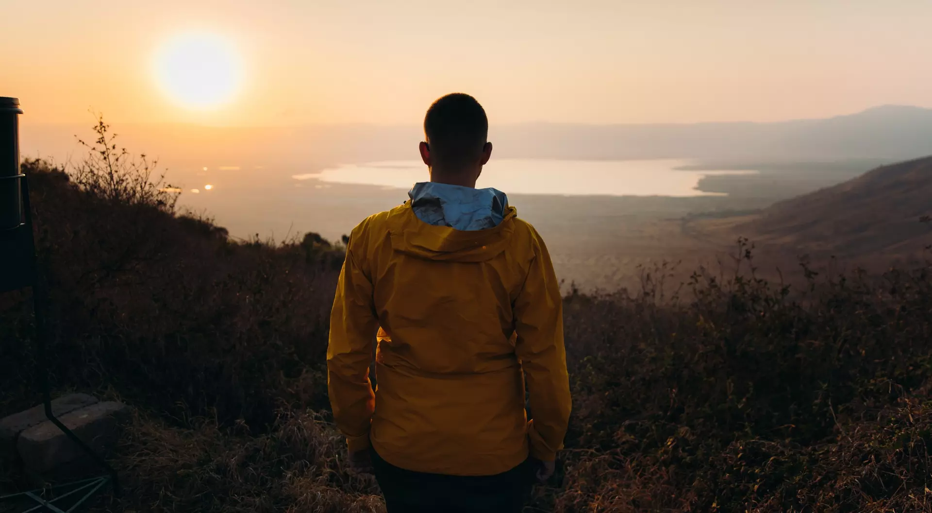 A person in a yellow jacket stands facing a sunset over a lake, surrounded by hills and vegetation.