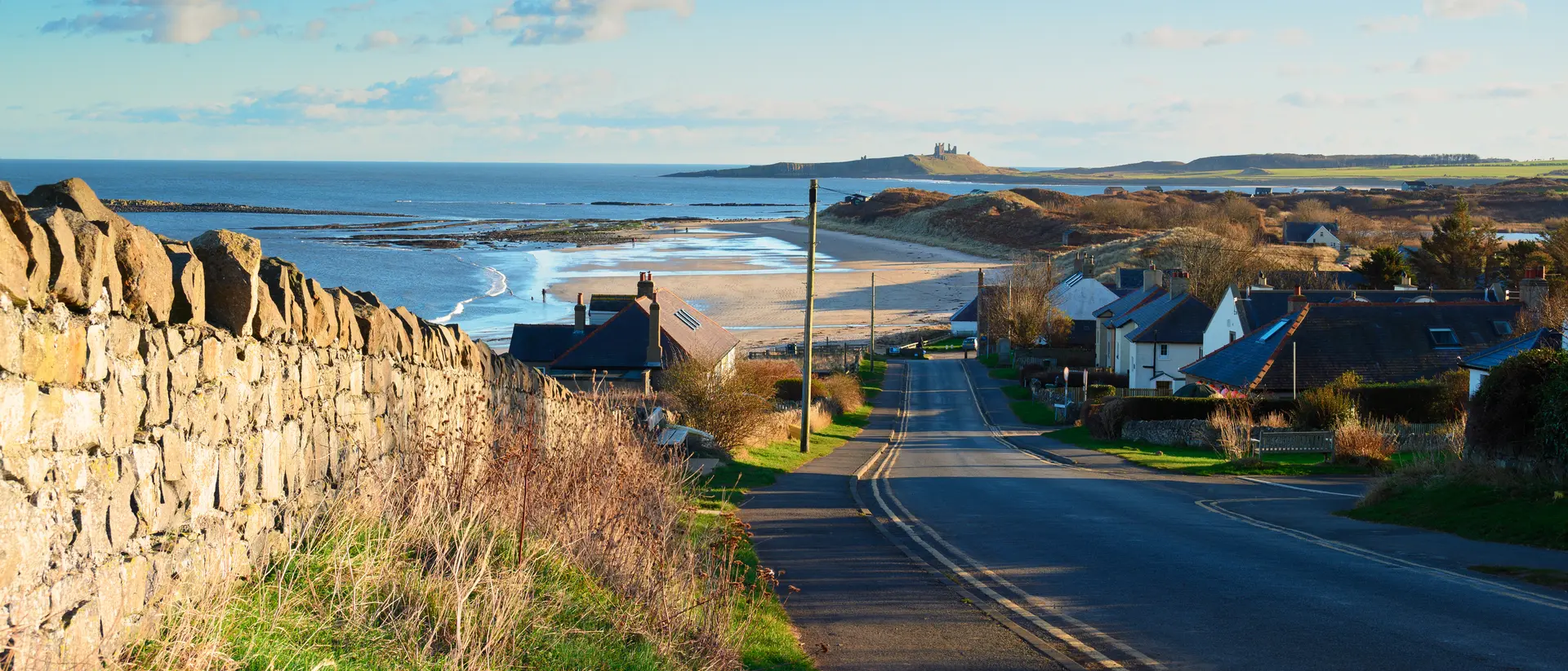 Scenic coastal view with a road, houses, and beach under a clear blue sky and distant hills.