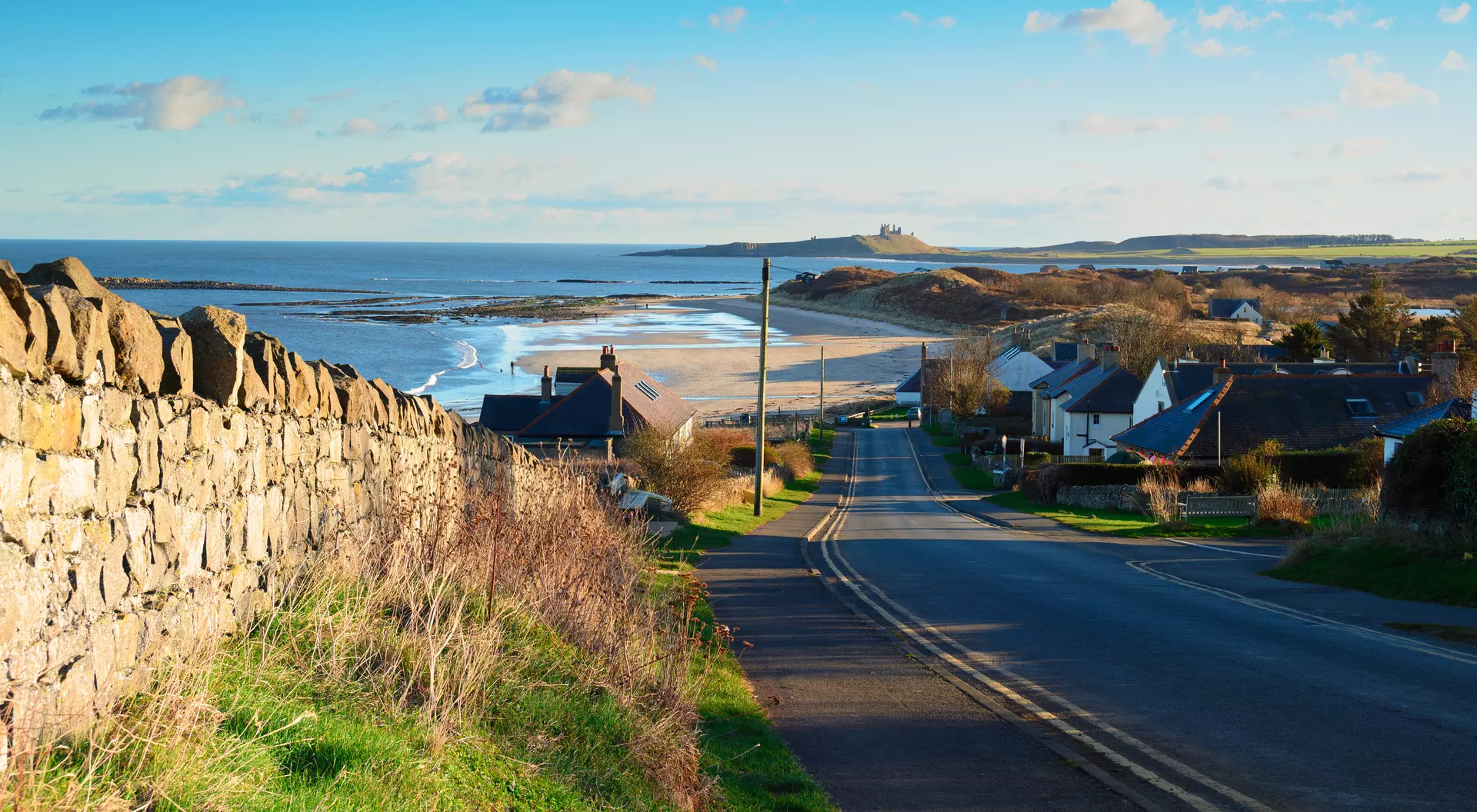 Scenic coastal view with a road, houses, and beach under a clear blue sky and distant hills.