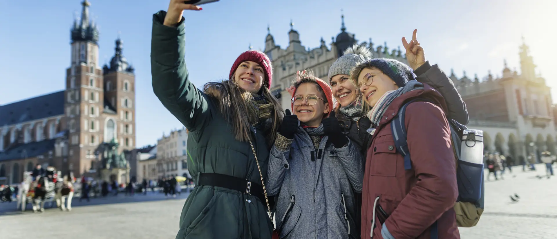 Four friends take a selfie in a sunny square, wearing winter coats and hats, with historic buildings in the background.