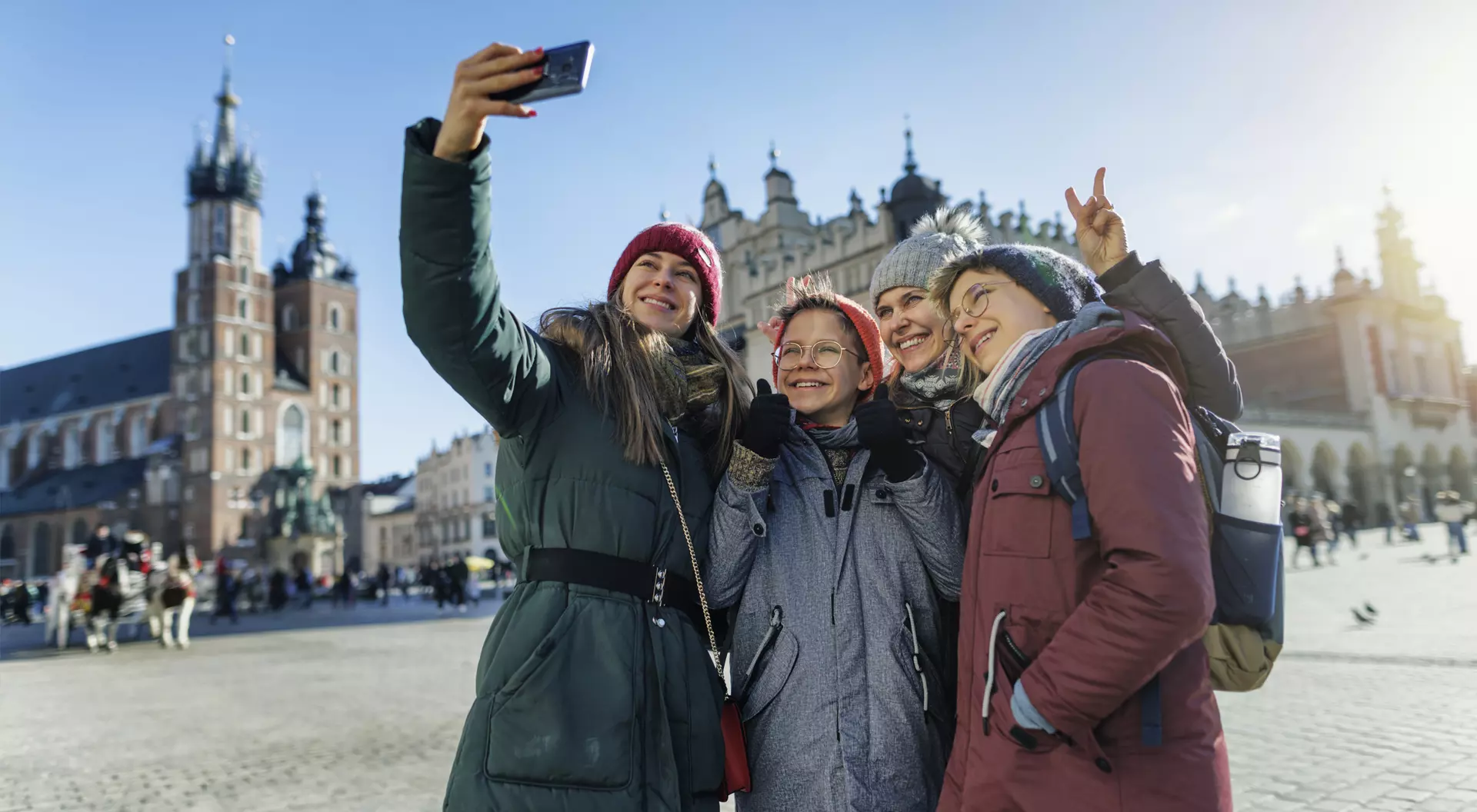 Four friends take a selfie in a sunny square, wearing winter coats and hats, with historic buildings in the background.