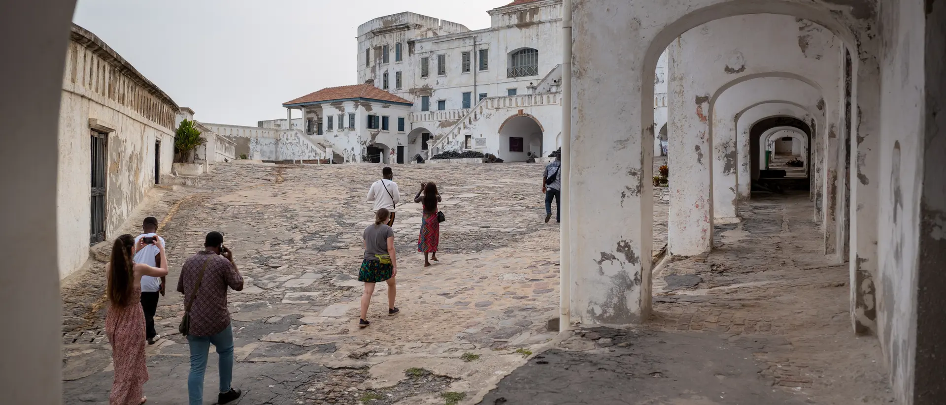 Group of people walking through an old, weathered courtyard with white buildings and arches in the background.