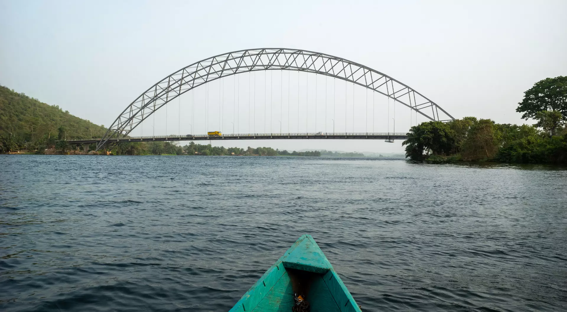 View from a boat on water towards a curved bridge spanning a river, surrounded by greenery on both banks.