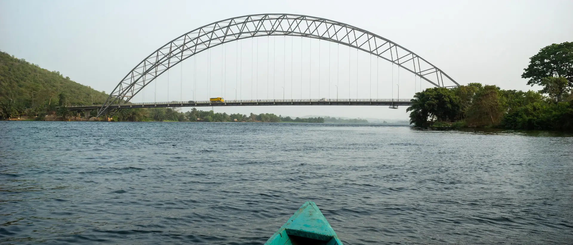 View from a boat on water towards a curved bridge spanning a river, surrounded by greenery on both banks.