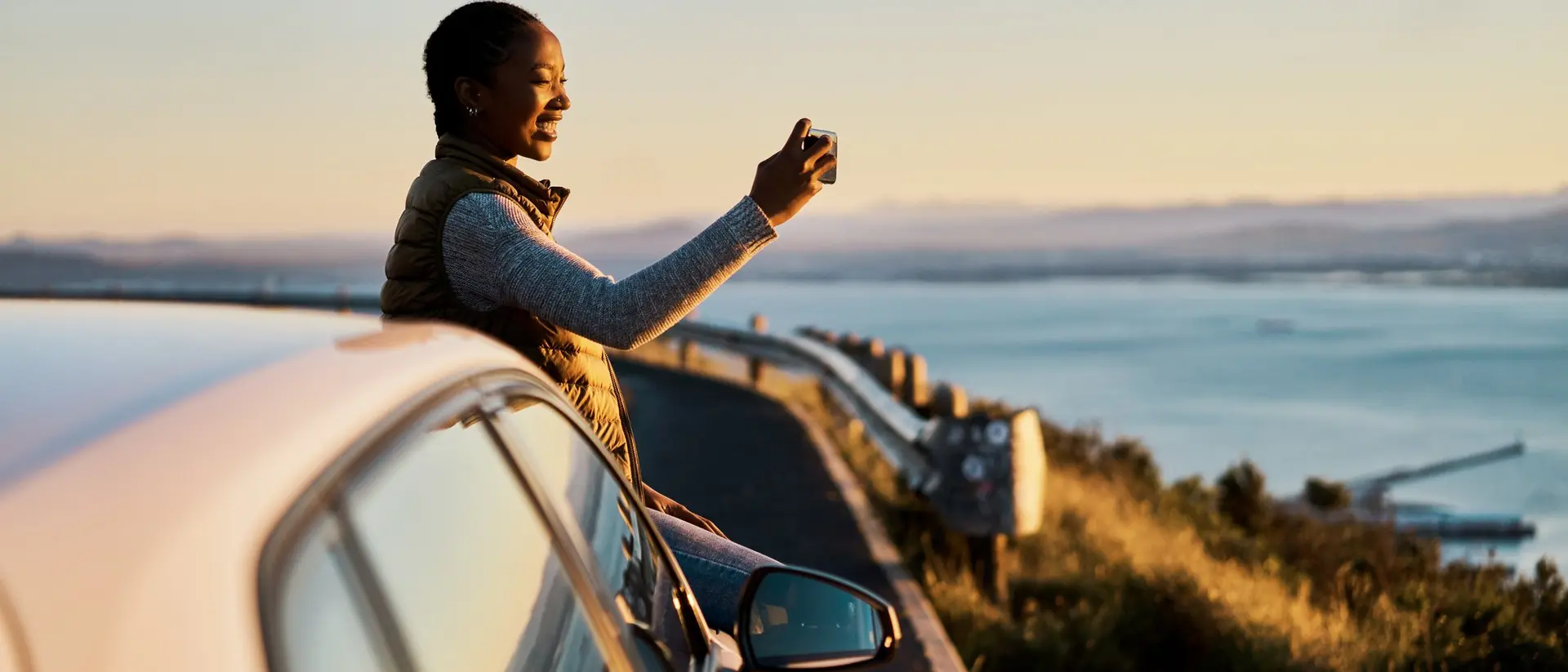 A person joyfully taking a selfie while sitting on a car by a scenic overlook at sunset.