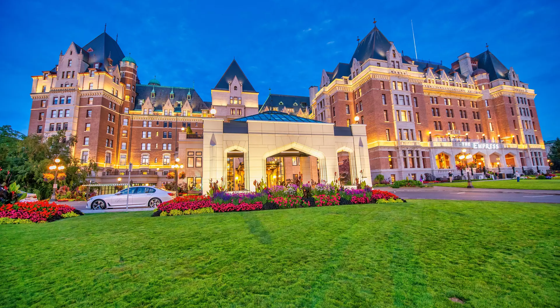 Historic hotel with a grand facade, vibrant gardens, and evening lights under a blue sky.