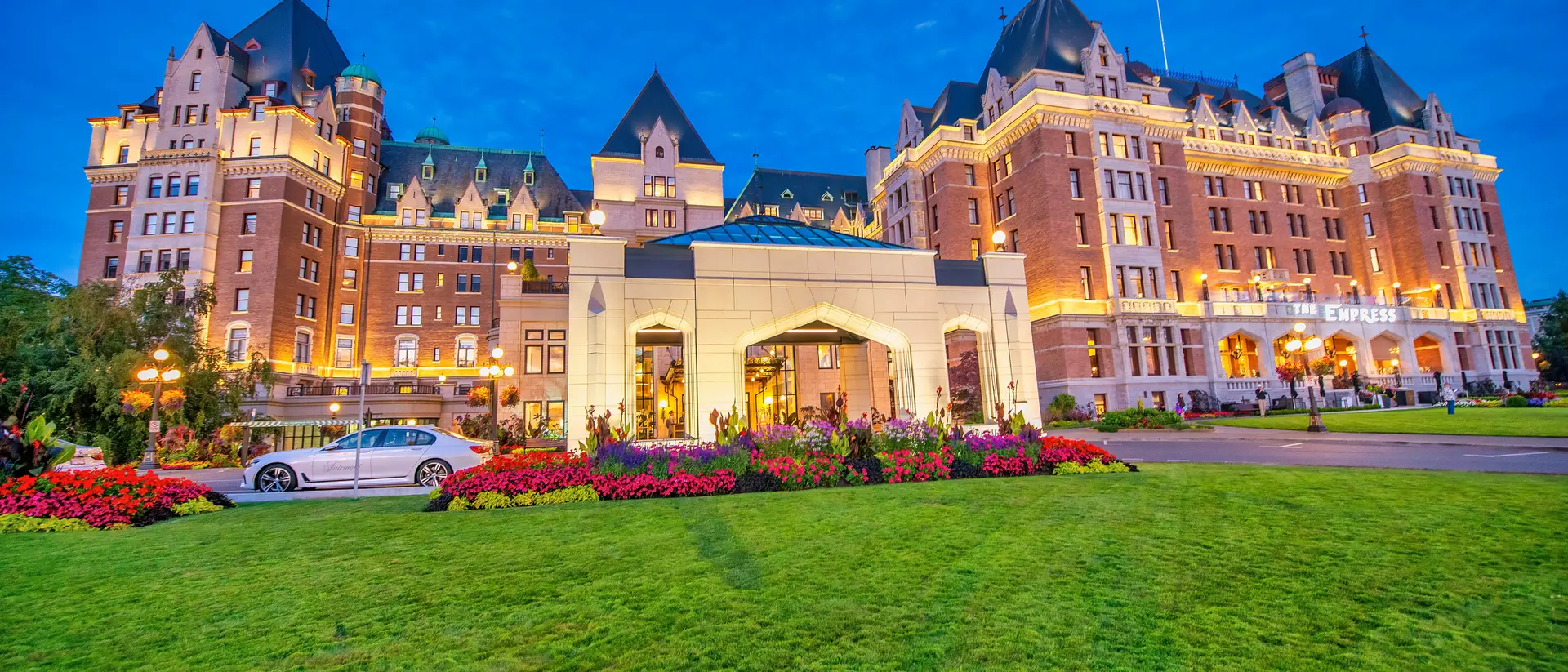 Historic hotel with a grand facade, vibrant gardens, and evening lights under a blue sky.