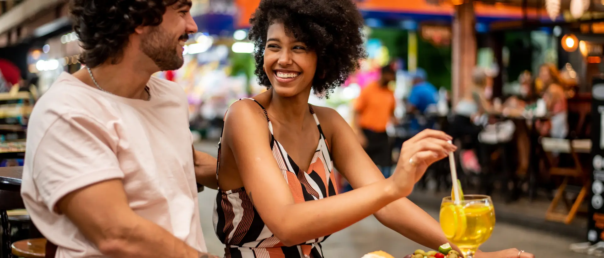A couple enjoys a meal at a lively outdoor restaurant, smiling and sharing food and drinks in a vibrant atmosphere.