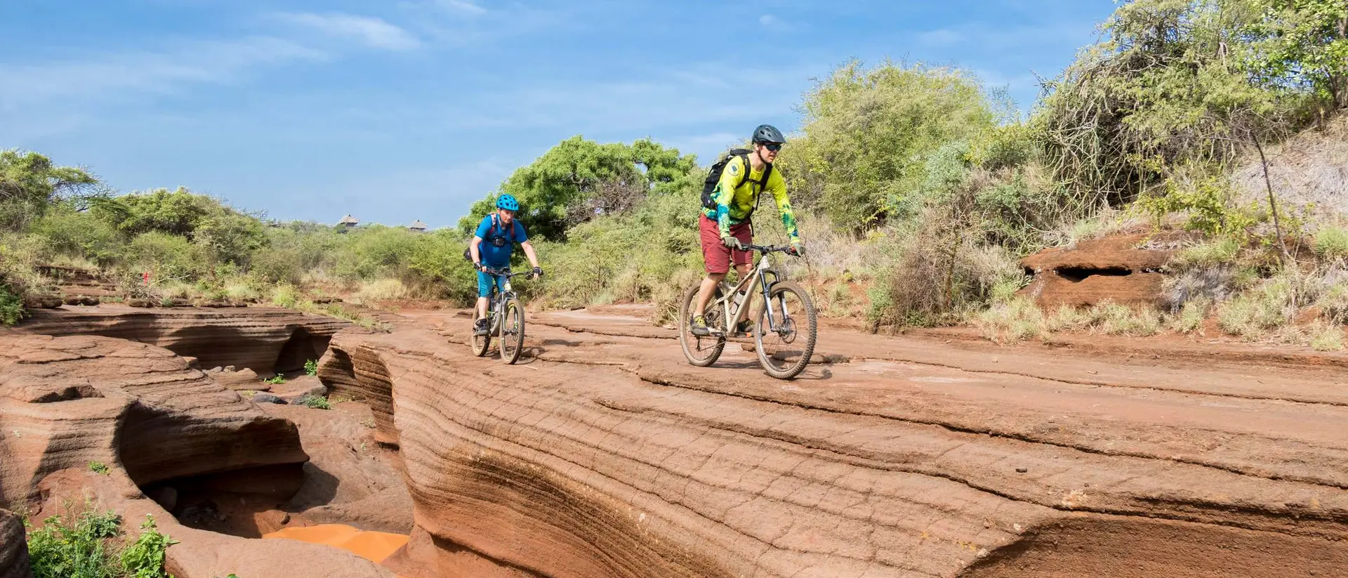 Two mountain bikers ride along a rocky, terraced landscape under a blue sky, surrounded by green vegetation.