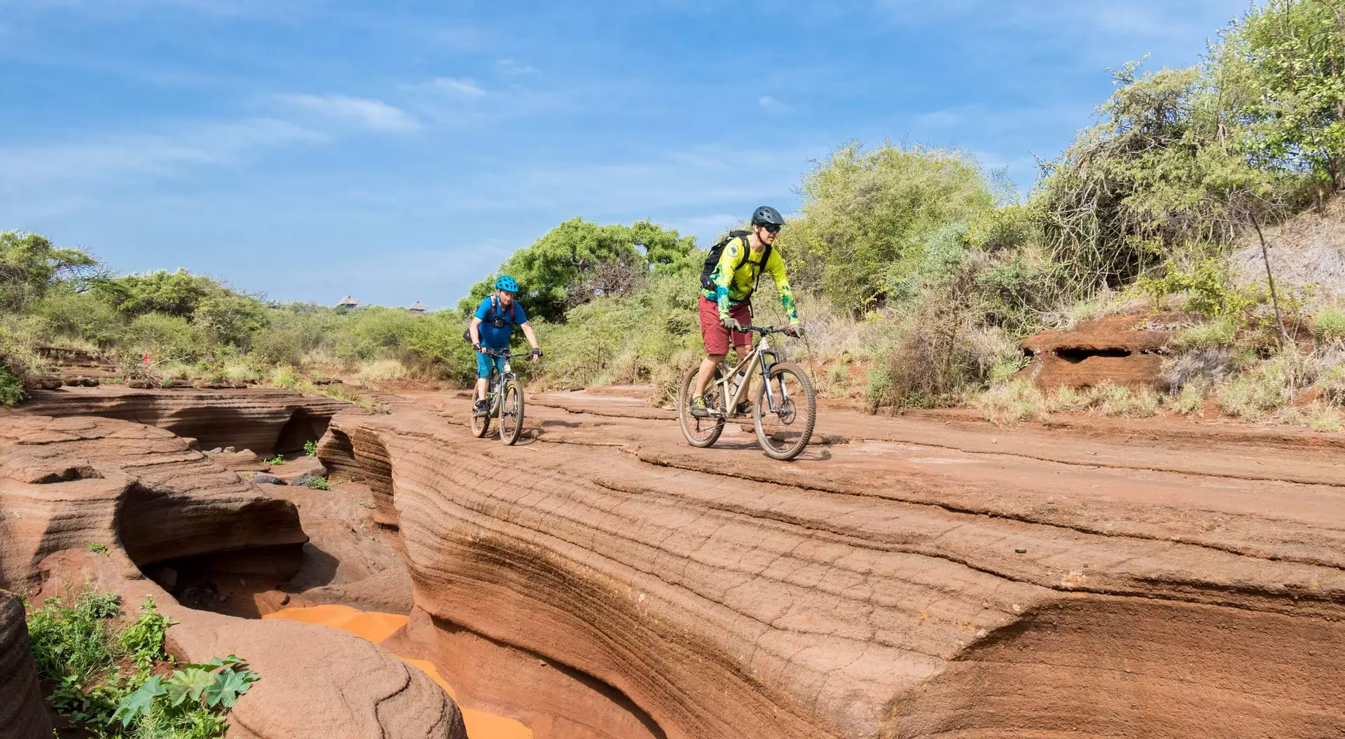 Two mountain bikers ride along a rocky, terraced landscape under a blue sky, surrounded by green vegetation.
