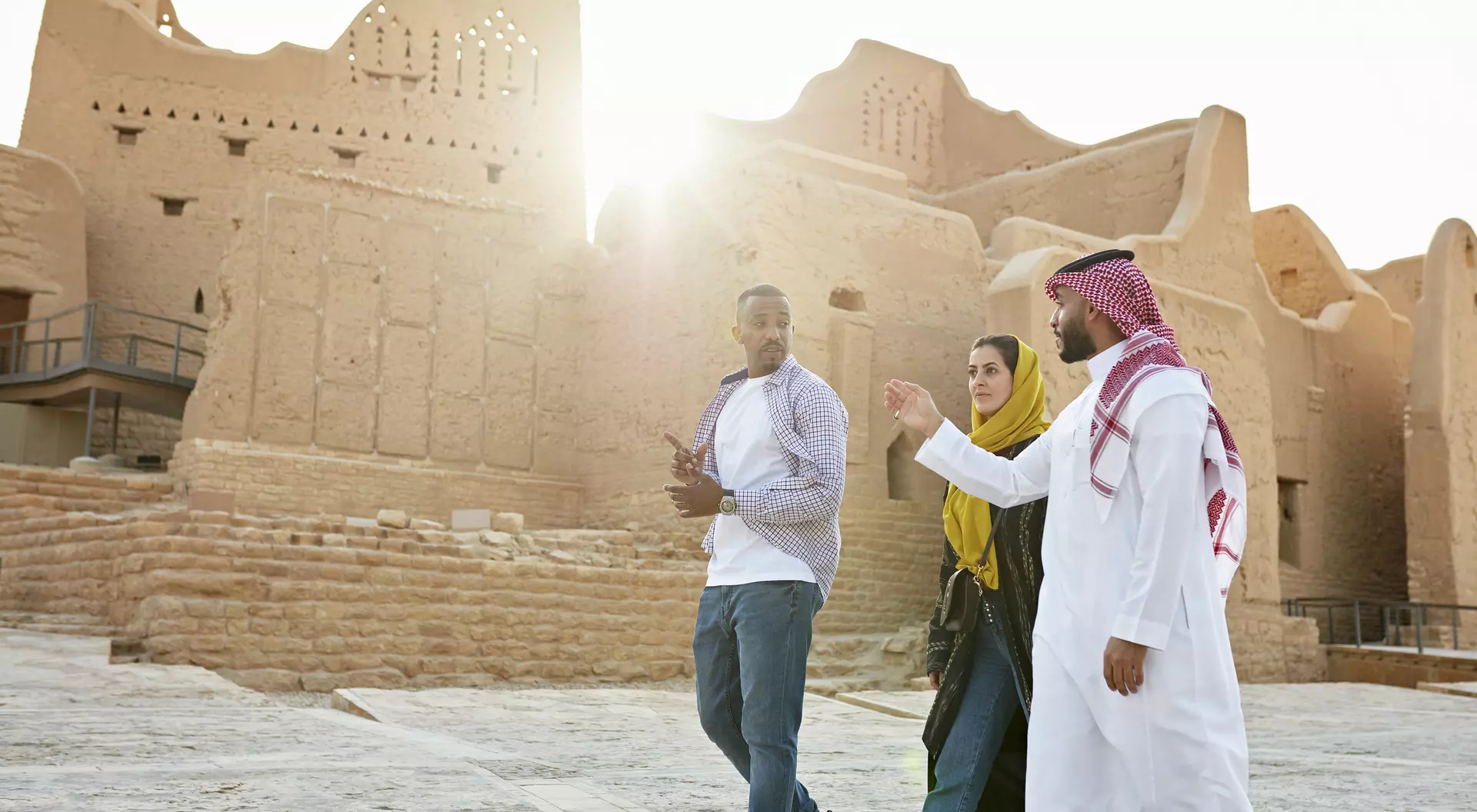 Three people walk together near historic mud-brick architecture, with sunlight shining in the background.