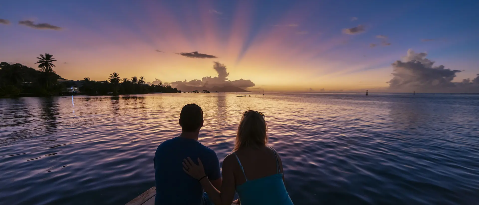 A couple sits on a dock at sunset, wading through calm water, with colorful rays of light illuminating the sky.