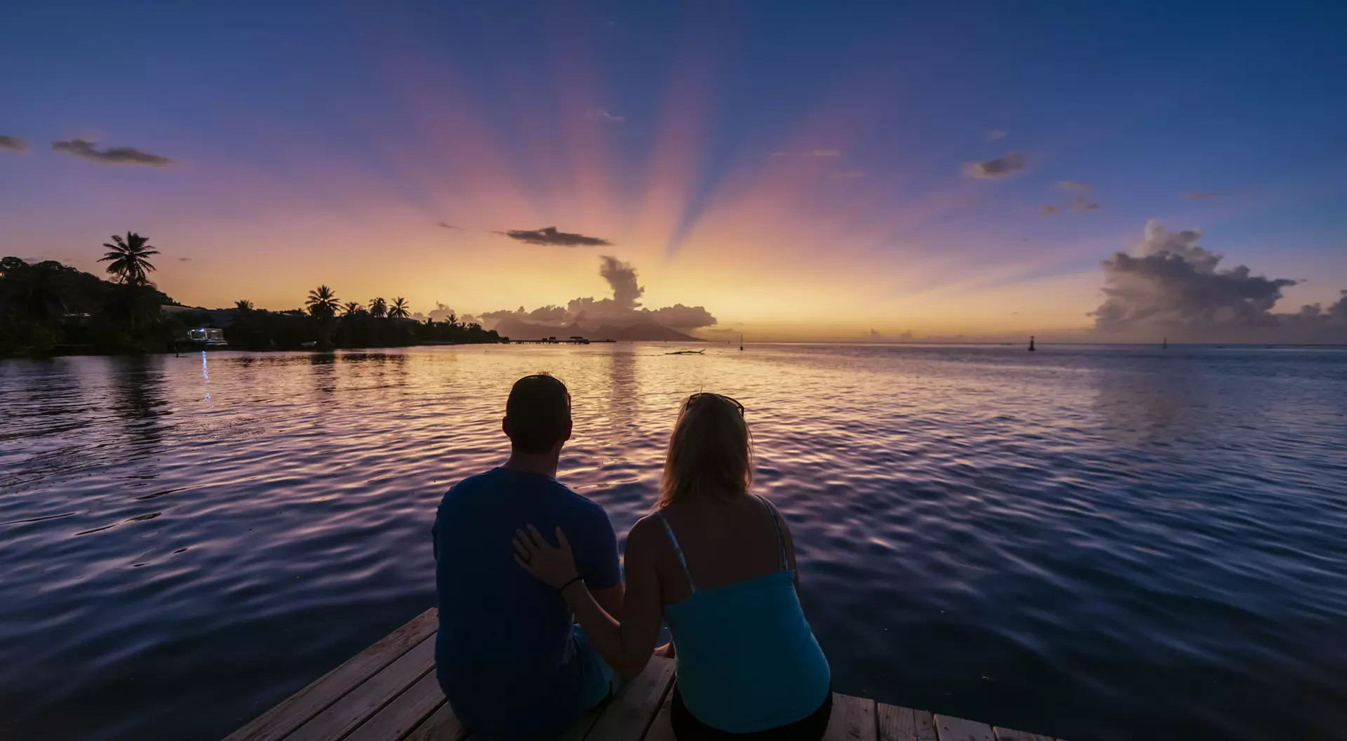 A couple sits on a dock at sunset, wading through calm water, with colorful rays of light illuminating the sky.