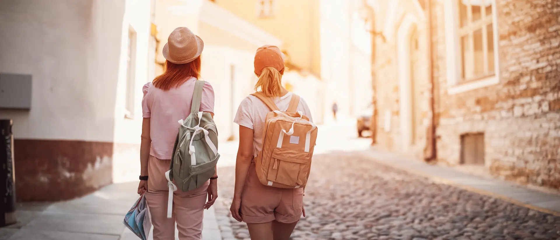 Two women walking down a cobblestone street, each wearing a backpack and enjoying a sunny day.