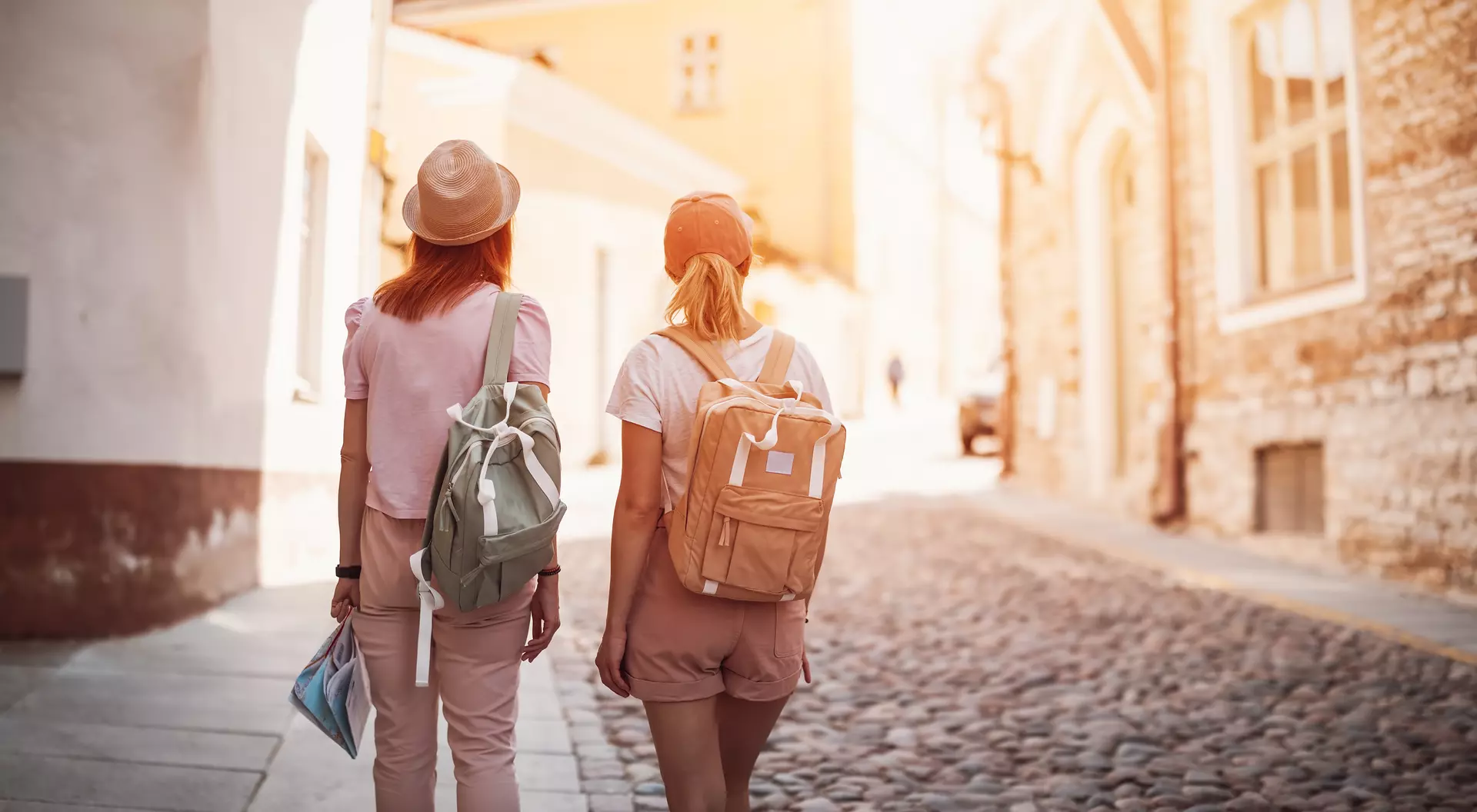 Two women walking down a cobblestone street, each wearing a backpack and enjoying a sunny day.