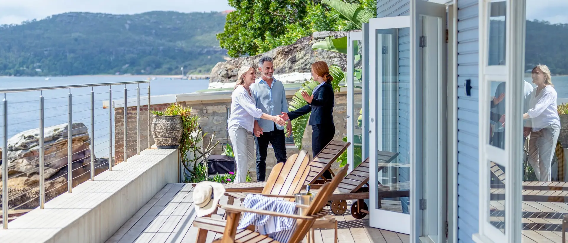 Three people shake hands on a sunny deck overlooking a scenic waterfront, with cozy chairs and plants nearby.