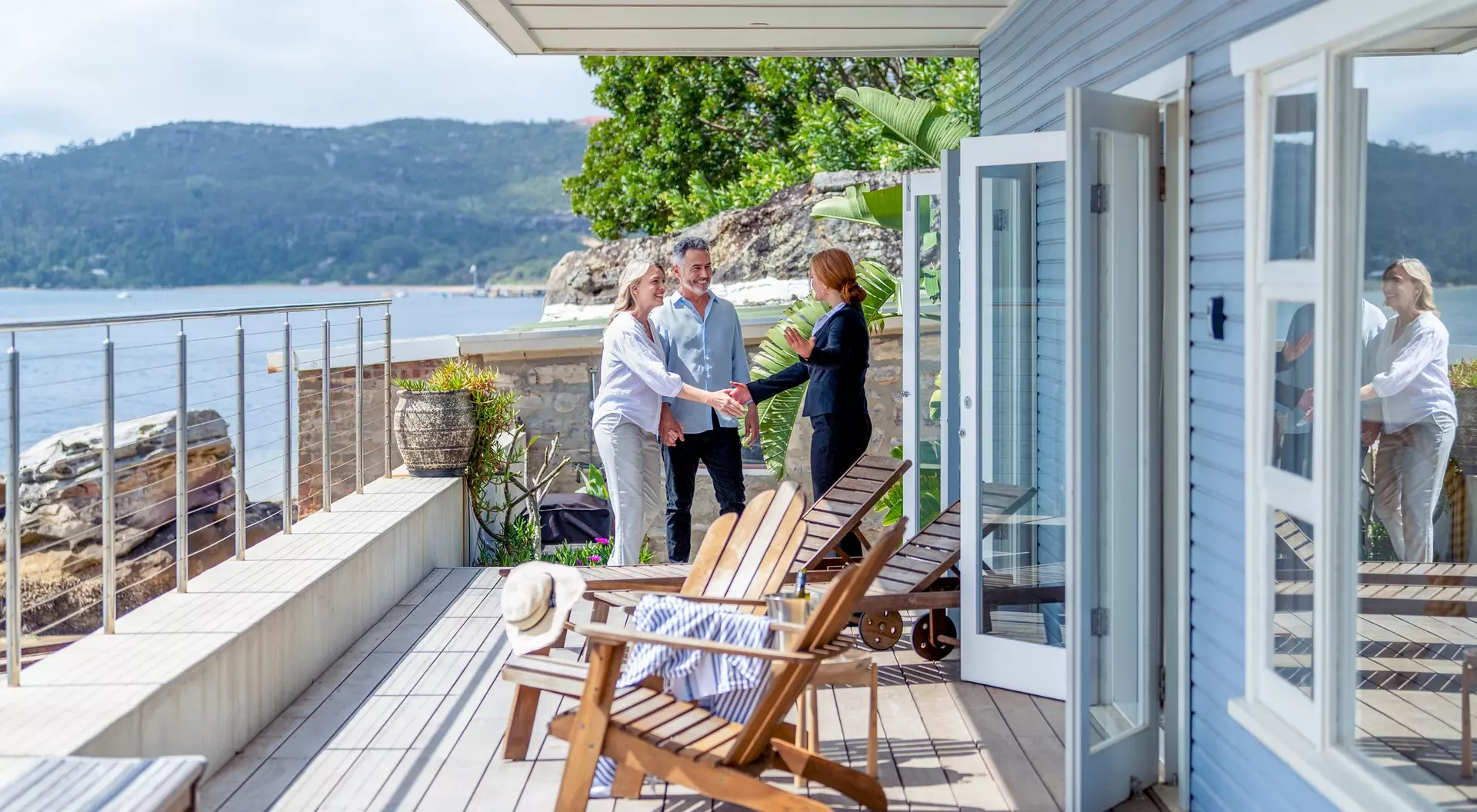 Three people shake hands on a sunny deck overlooking a scenic waterfront, with cozy chairs and plants nearby.
