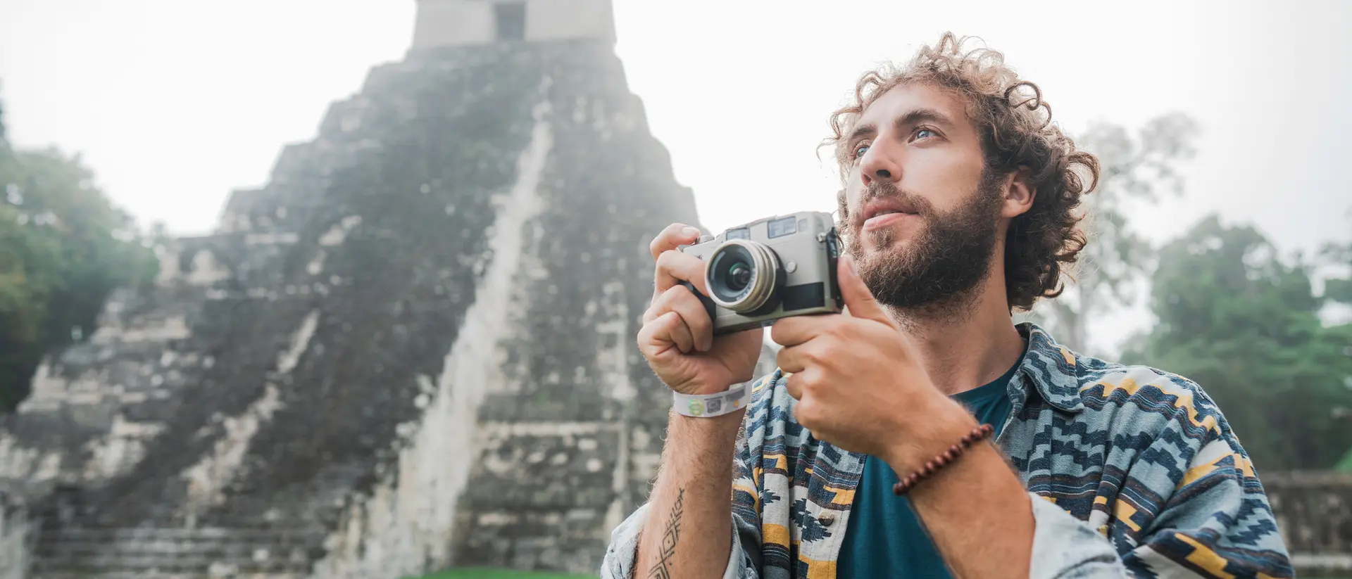 A man with curly hair holds a camera, standing in front of a Mayan pyramid in a misty landscape.