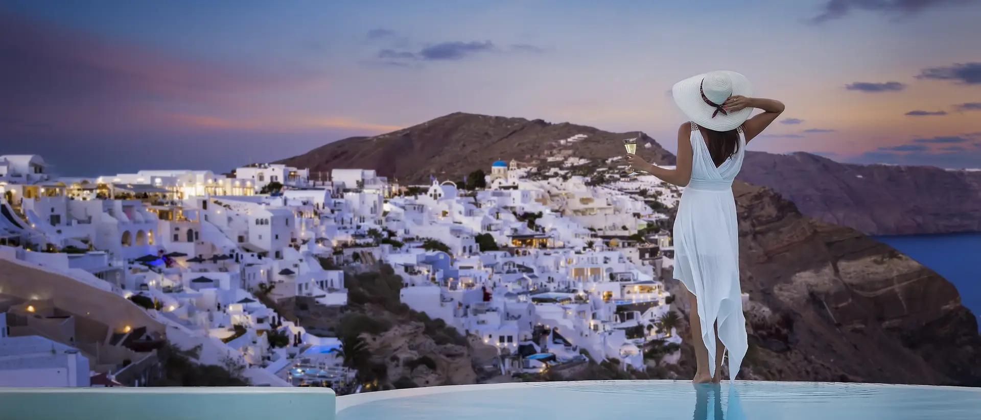 A stands by the swimming pool and enjoys the sunset view over the illuminated village of Oia