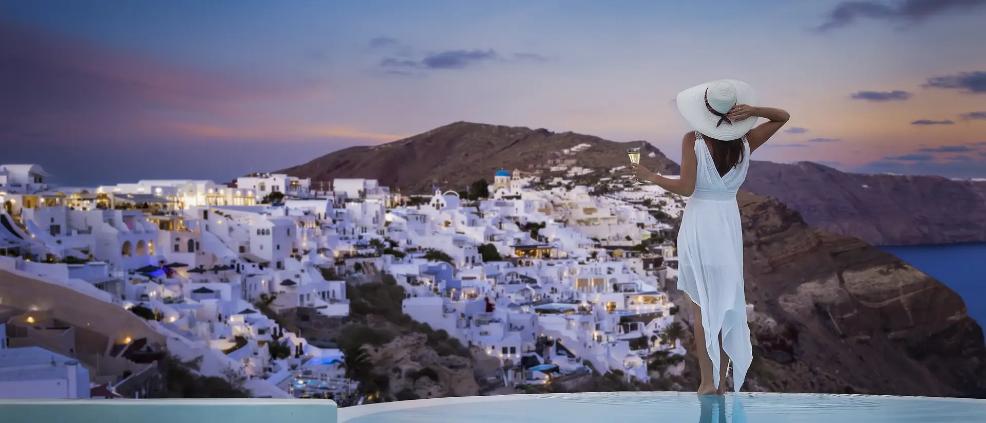 A stands by the swimming pool and enjoys the sunset view over the illuminated village of Oia