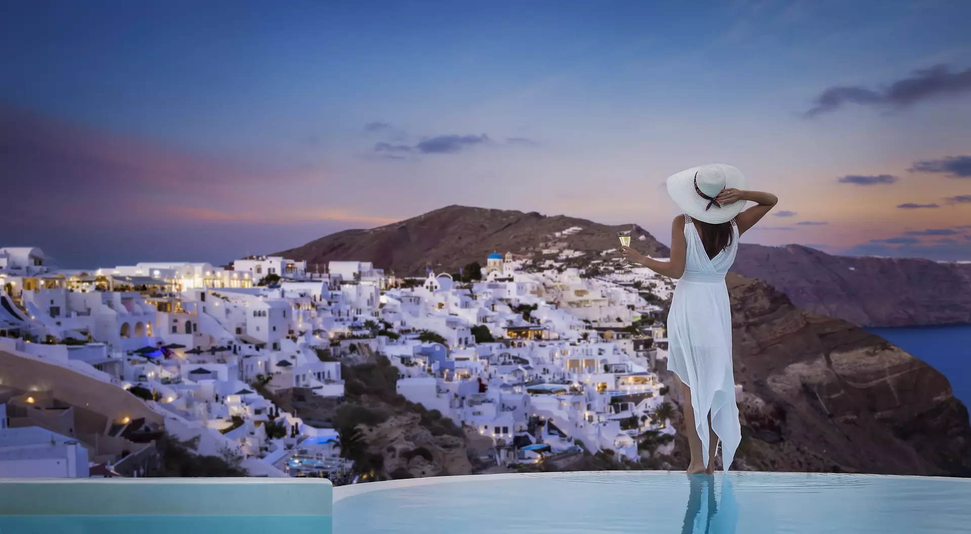 A stands by the swimming pool and enjoys the sunset view over the illuminated village of Oia