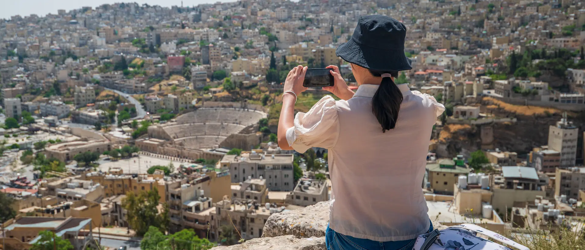 A person in a hat takes a photo of a city view, showcasing hills and buildings in the background.