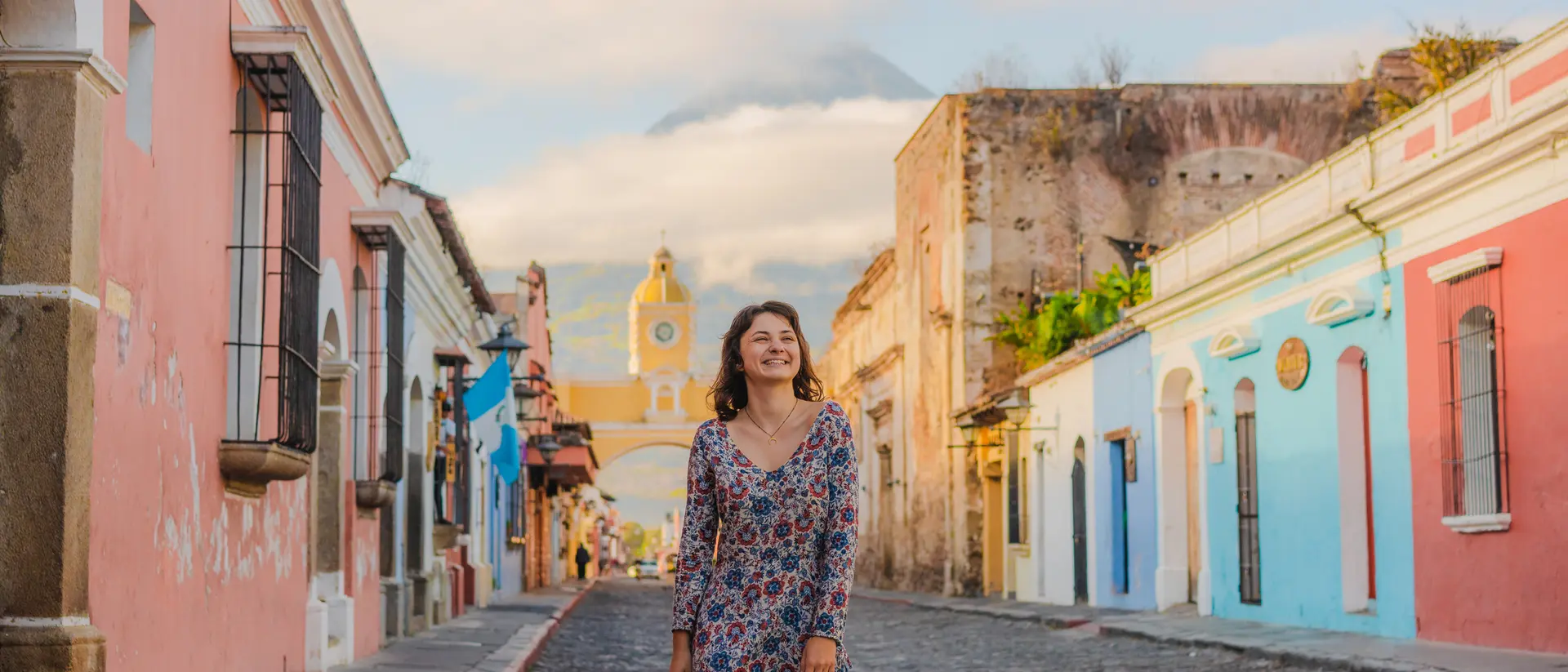 A woman stands on a cobblestone street lined with colorful buildings, with a mountain in the background under a clear sky.