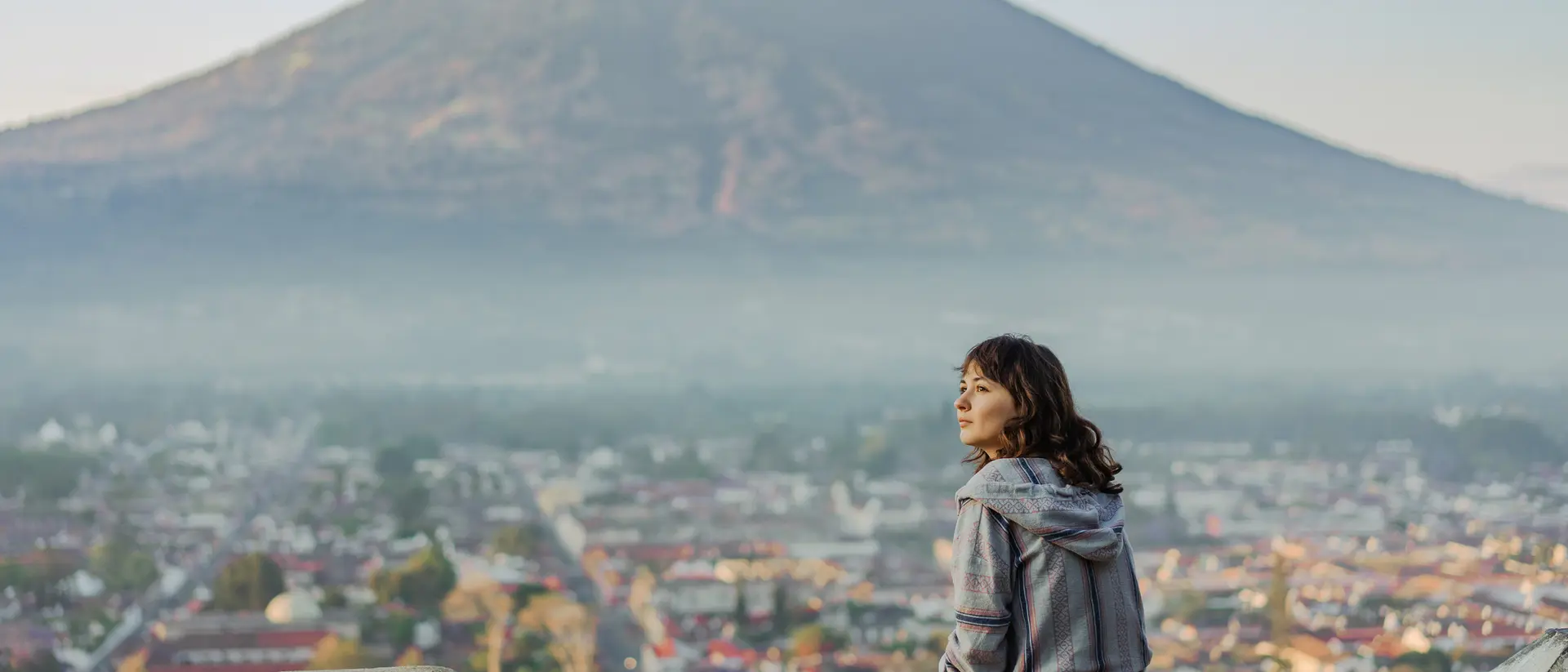 A person sits on a stone wall, overlooking a valley with a volcano in the background, surrounded by misty mountains.