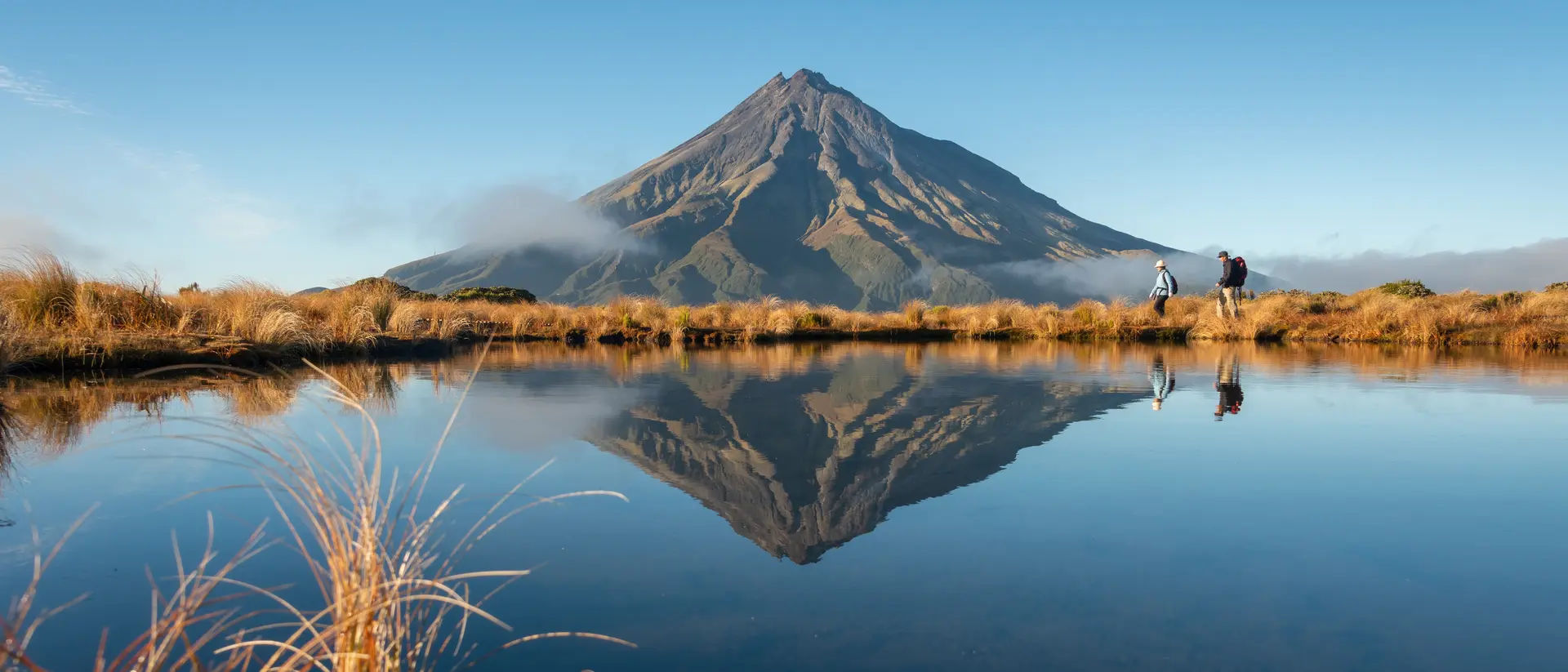 Two people hiking with the mountain reflected in the tarn in early morning sunshine
