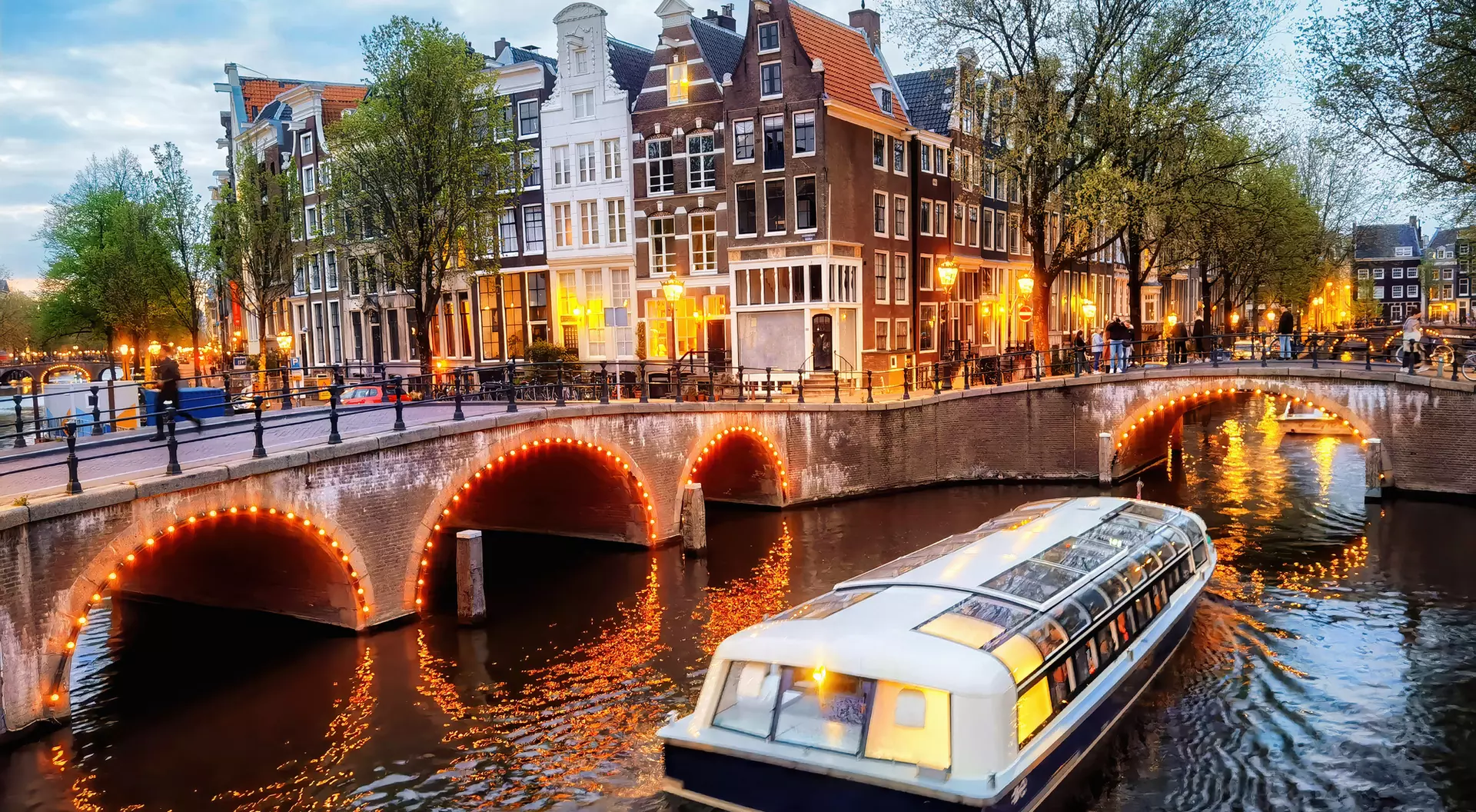 A scenic view of a canal in Amsterdam with a boat and historic buildings lit up at dusk.