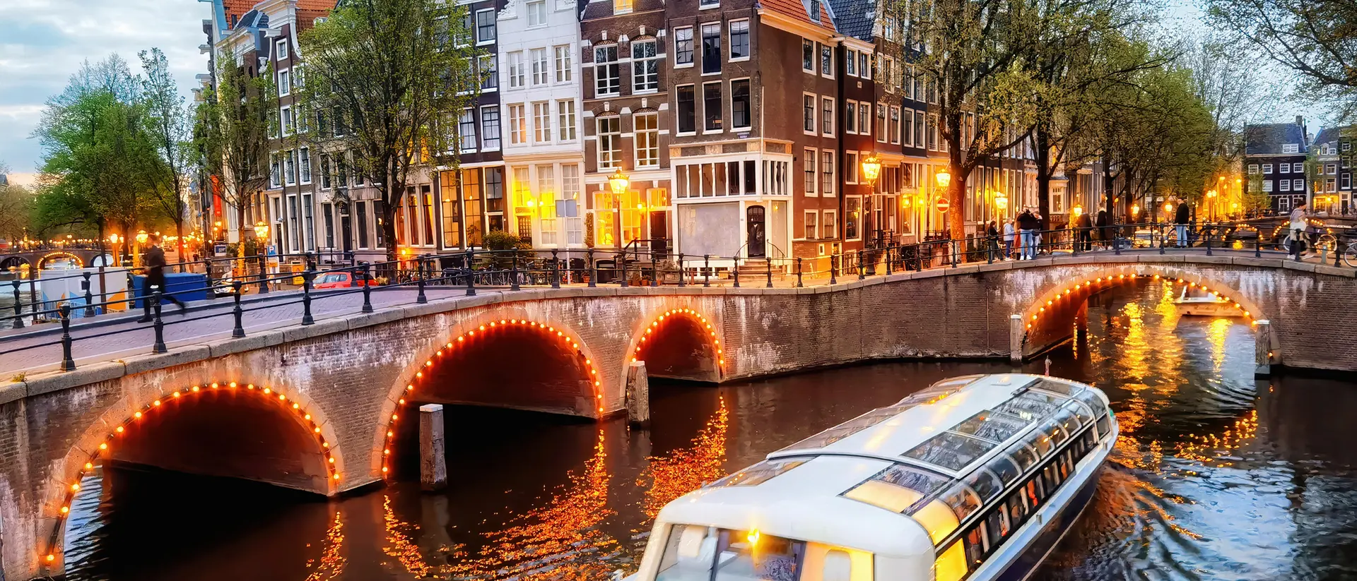 A scenic view of a canal in Amsterdam with a boat and historic buildings lit up at dusk.