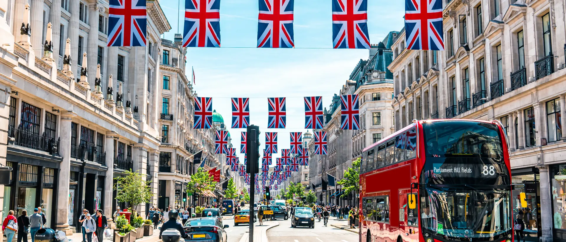 A busy street lined with shops, featuring red double-decker buses and decorative Union Jack flags overhead.