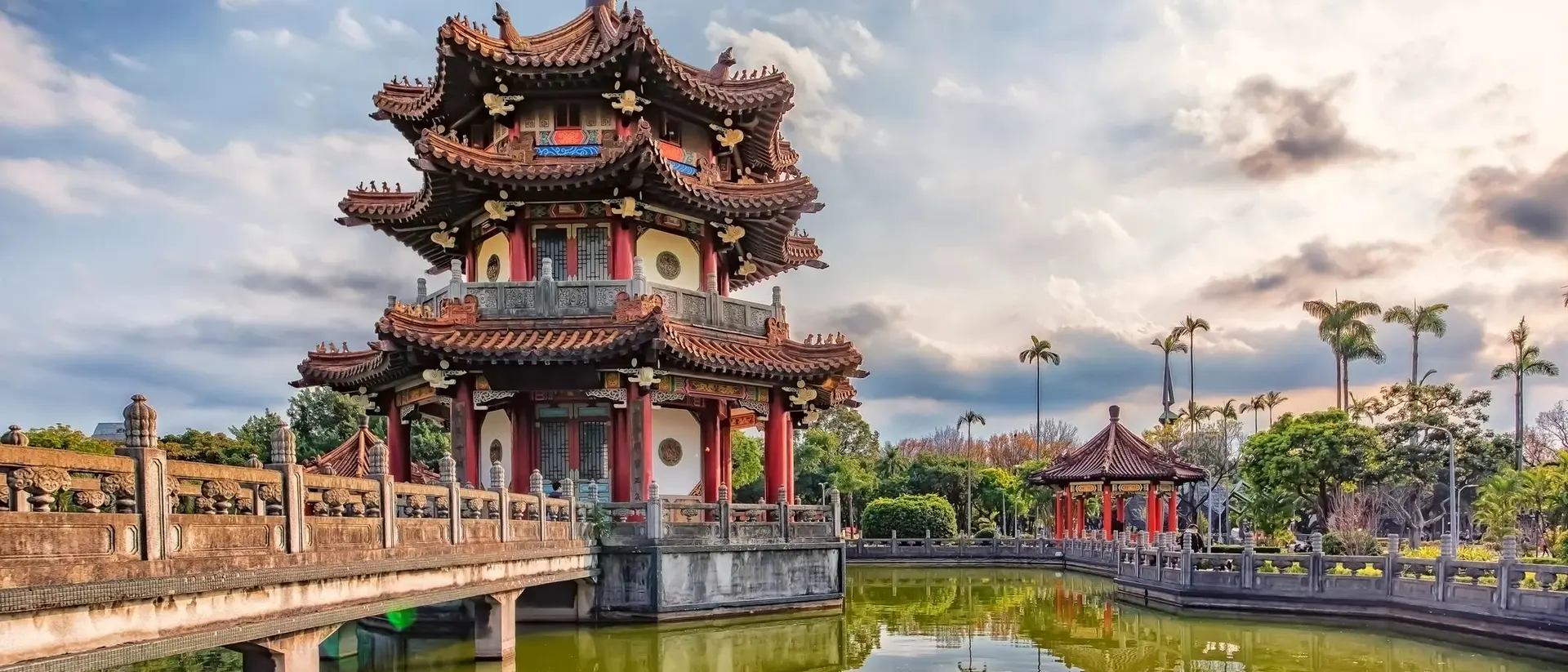 A traditional pagoda beside a tranquil pond, surrounded by lush greenery and a cloudy sky.