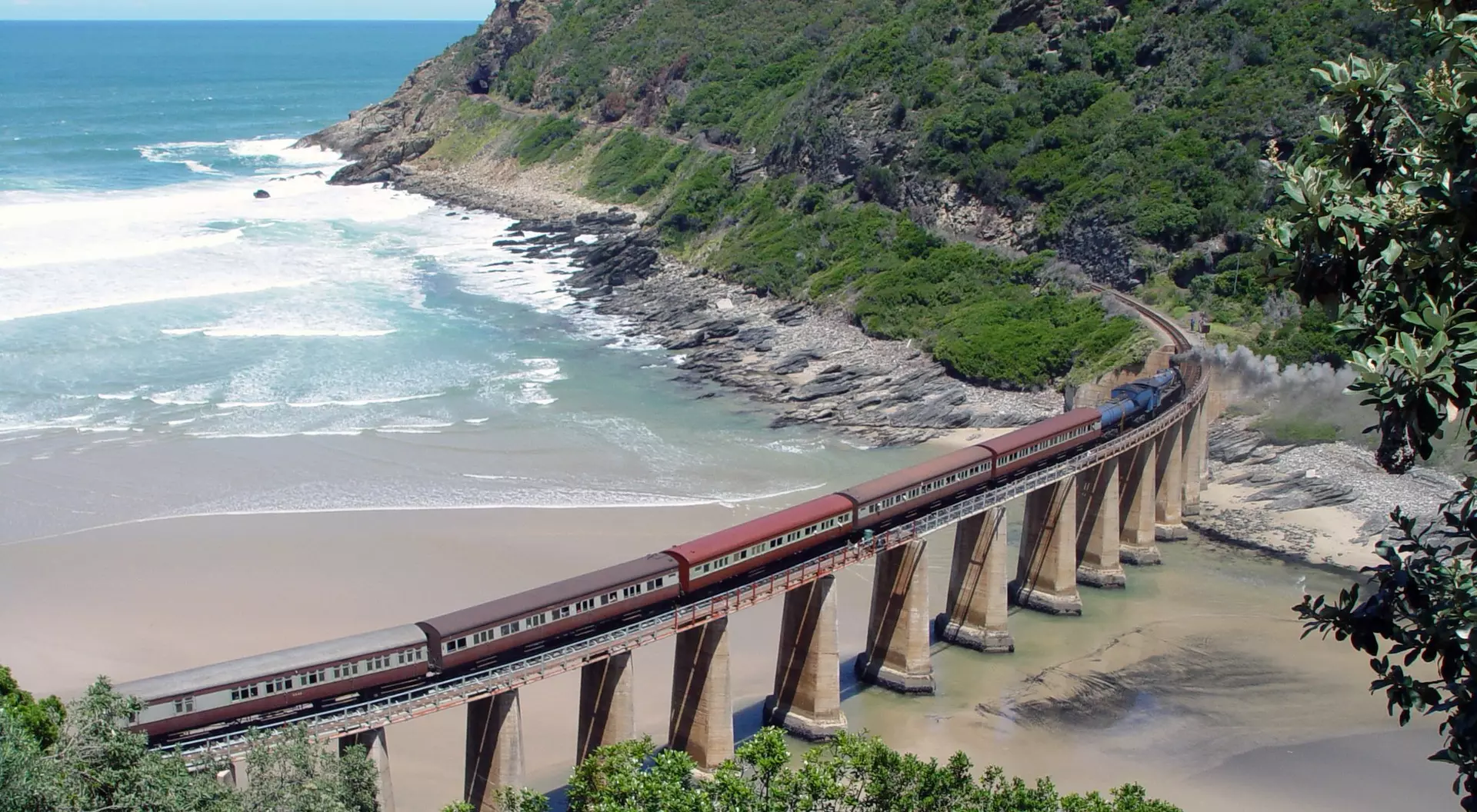 A scenic view of a train traversing a coastal railway bridge surrounded by lush greenery and the ocean.