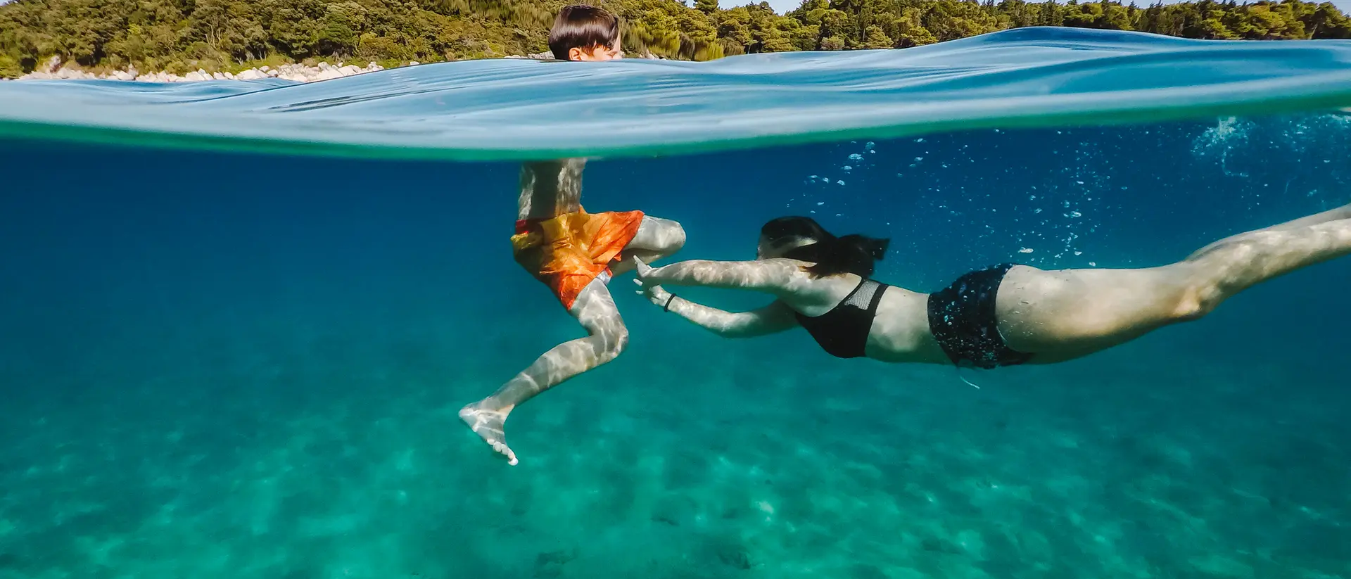 A child in swim trunks plays in the water with a person in a bikini, both enjoying a sunny day in a clear, blue sea.