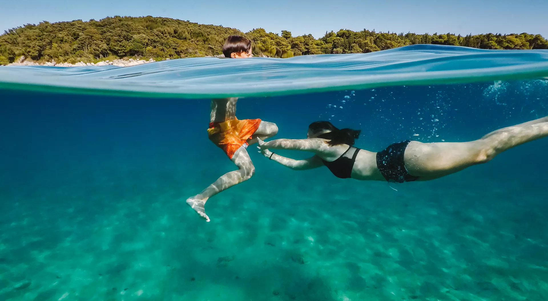A child in swim trunks plays in the water with a person in a bikini, both enjoying a sunny day in a clear, blue sea.