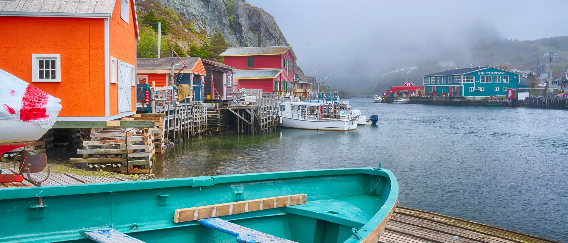 Colorful buildings line a foggy harbor with boats and a wooden dock, creating a tranquil coastal scene.