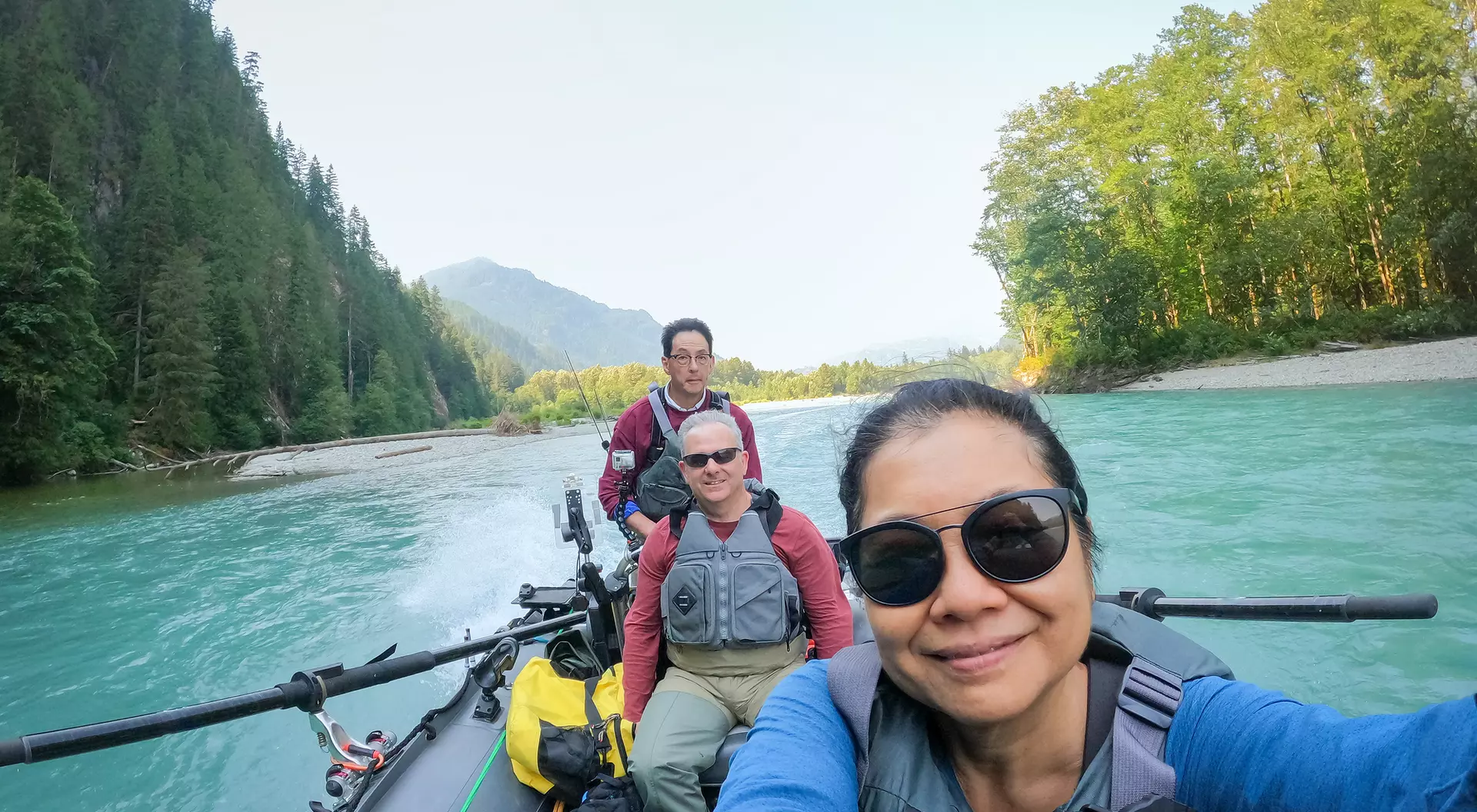 Three people in a boat on a river surrounded by trees and mountains, with one person taking a selfie.