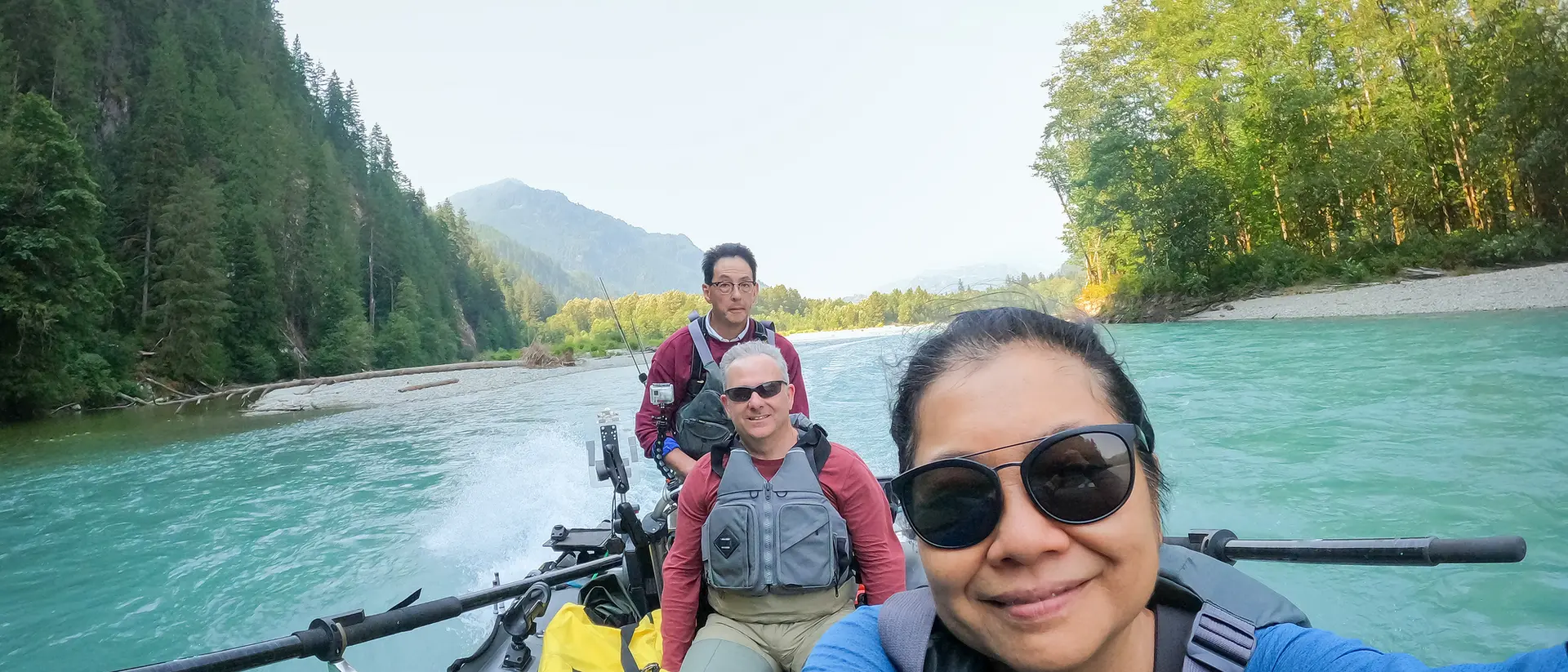 Three people in a boat on a river surrounded by trees and mountains, with one person taking a selfie.