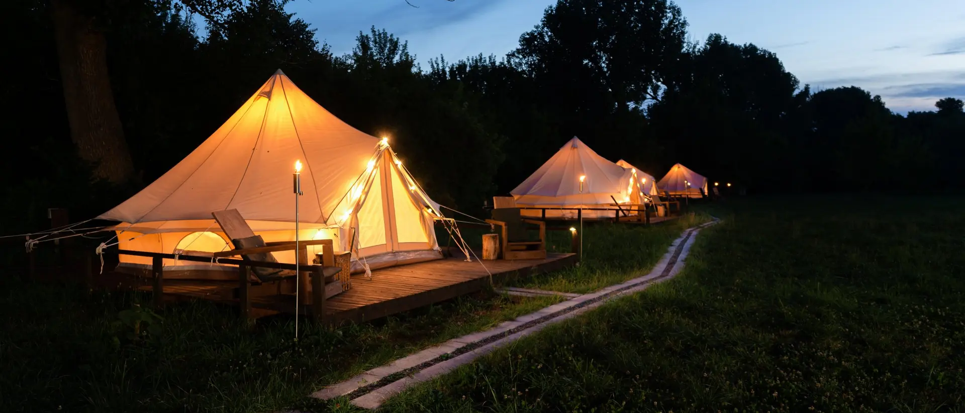 Tented accommodations glow warmly at dusk, surrounded by grass and trees, with a path leading through the campsite.