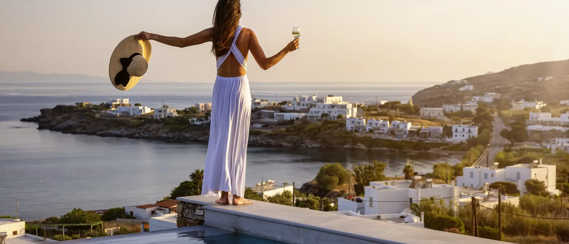 A woman in a white dress holds a drink and a large hat, standing by a pool with a scenic coastal view at sunset.