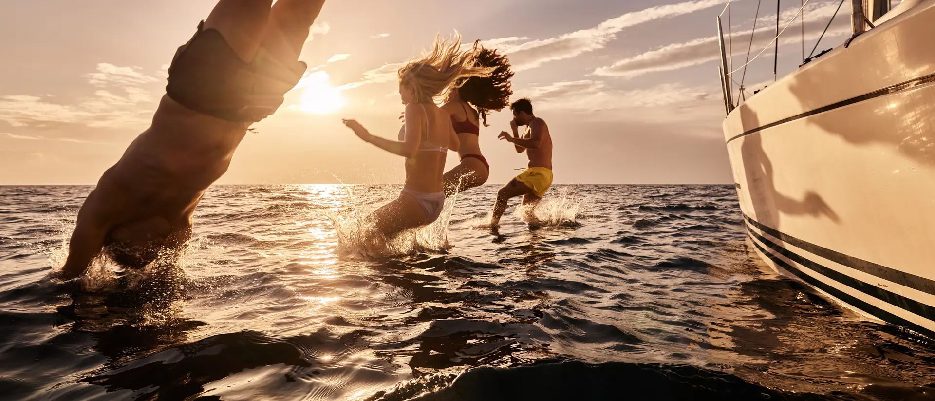 Three people jump into the ocean from a yacht at sunset, creating splashes in the water.