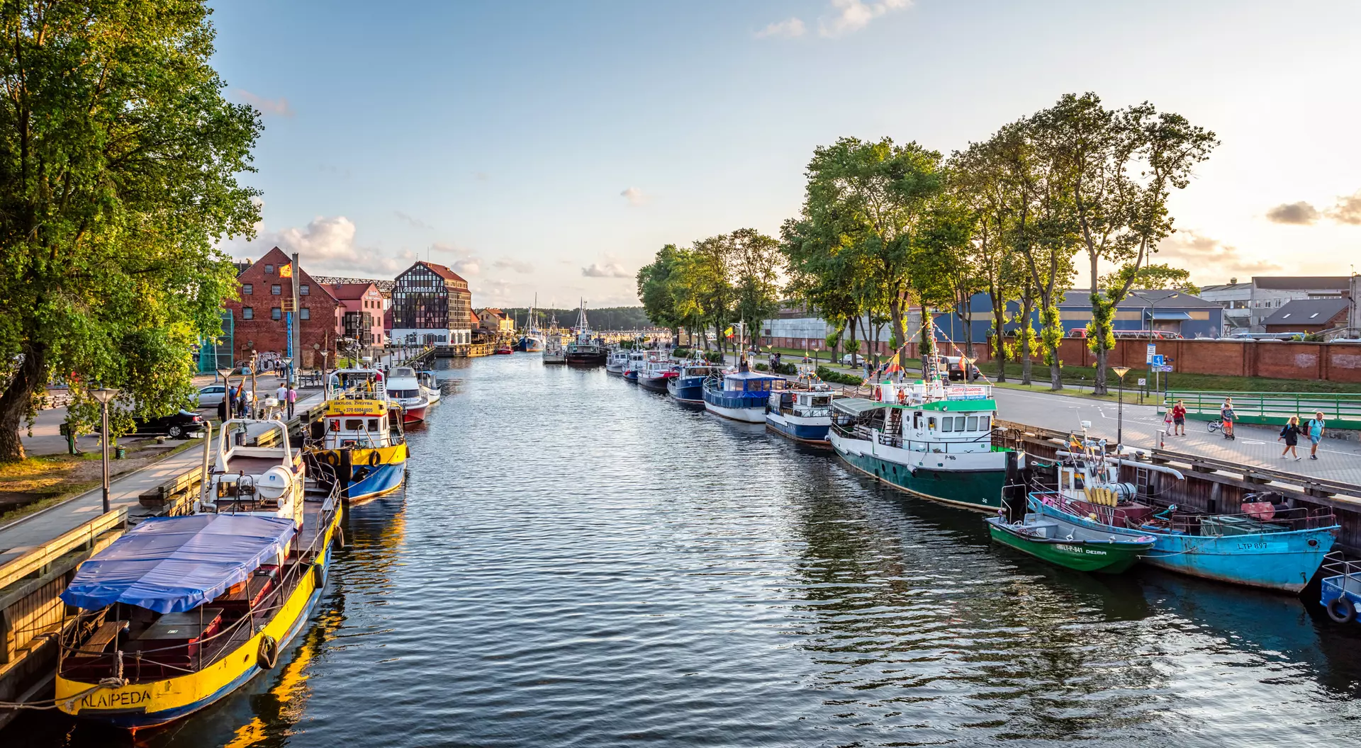 Colorful boats lined up along a calm river with trees and buildings in the background during sunset.