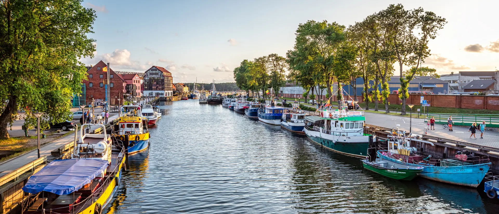 Colorful boats lined up along a calm river with trees and buildings in the background during sunset.