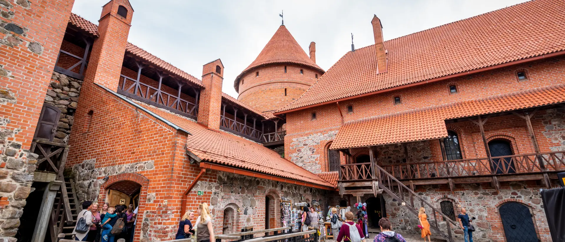 Visitors explore a historic castle courtyard with red brick buildings and a tower under a cloudy sky.